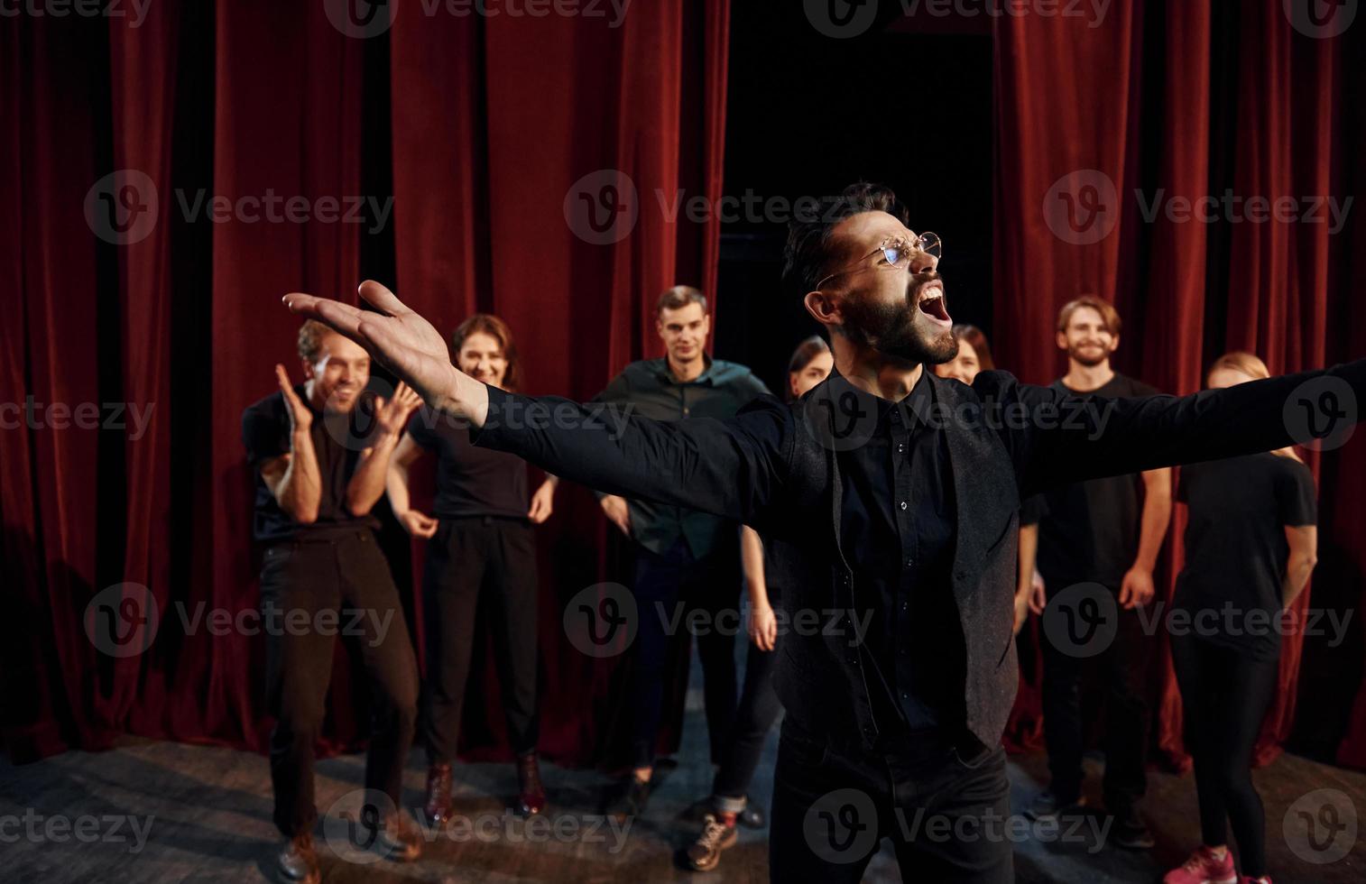 chico expresionable practicando su papel. grupo de actores con ropa de color oscuro ensayando en el teatro foto