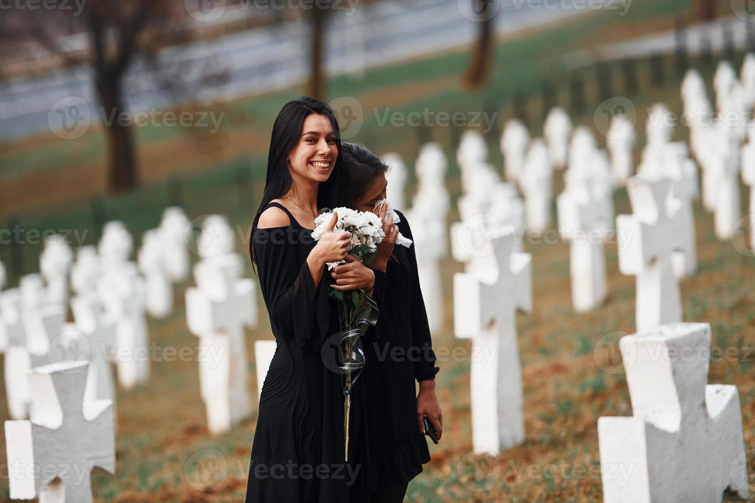 Girl smiling for no reason. Two young women in black clothes visiting cemetery with many white crosses. Conception of funeral and death photo