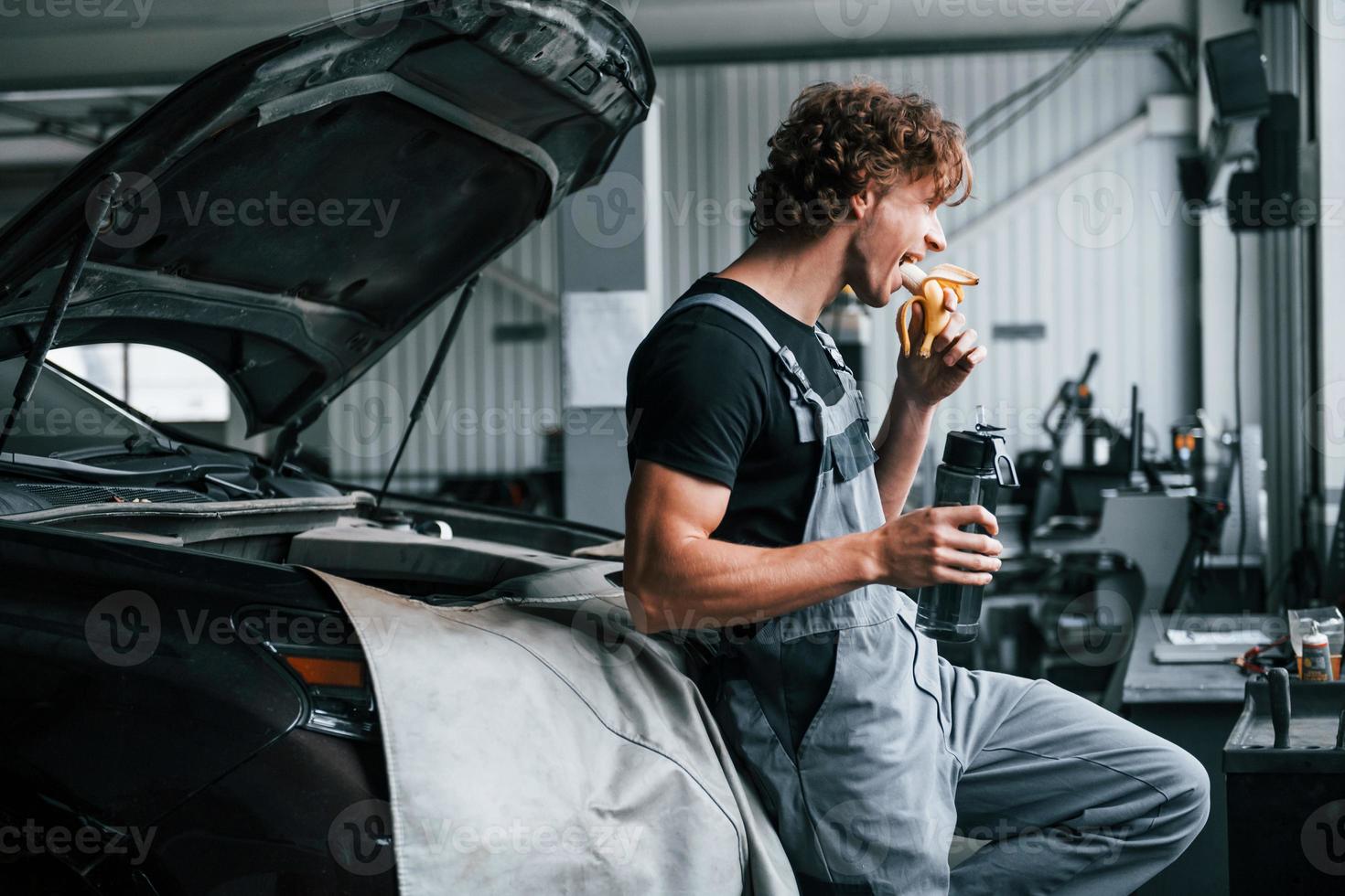 Takes a break. Eats banana and drinks water. Adult man in grey colored uniform works in the automobile salon photo