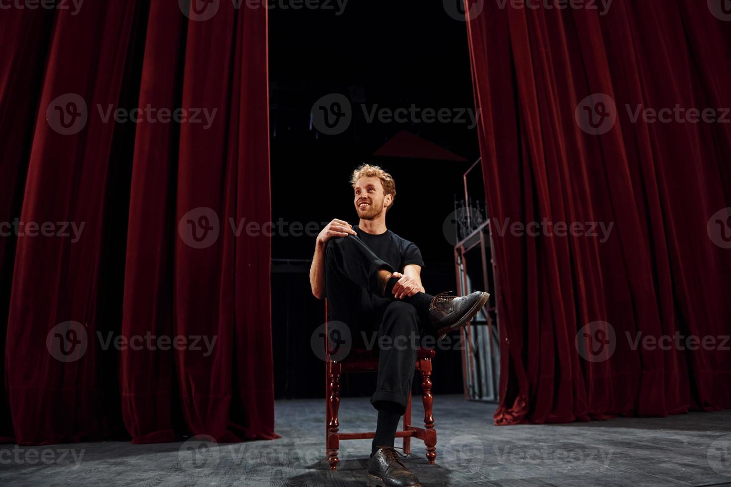 joven positivo sentado en la silla entre las cortinas en el escenario del teatro foto