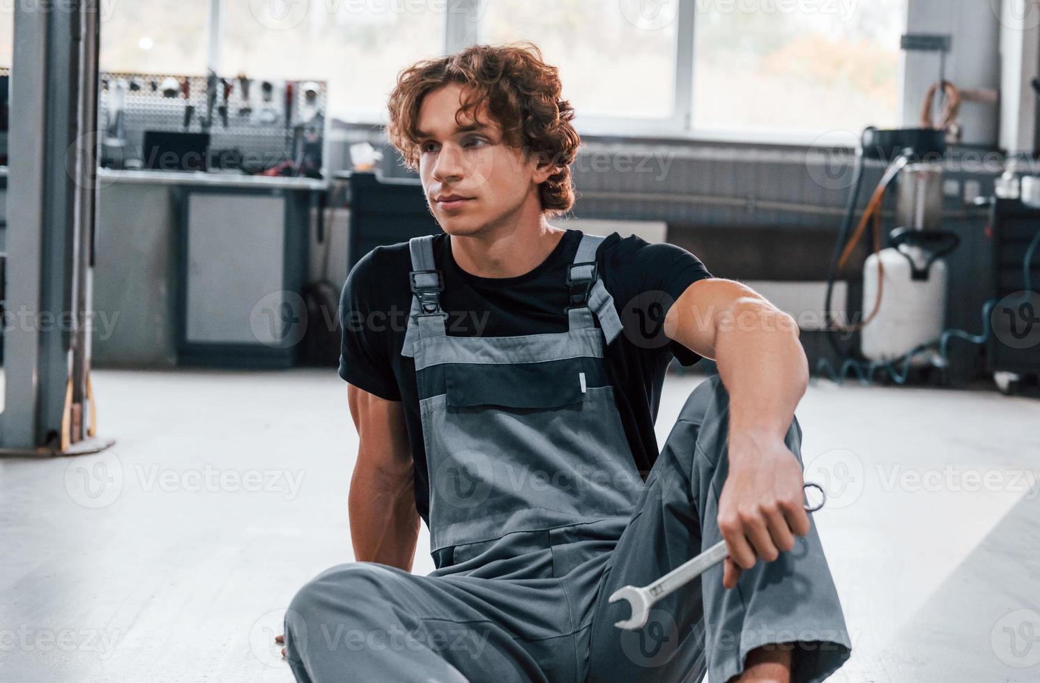 With tool in hand. Adult man in grey colored uniform works in the automobile salon photo