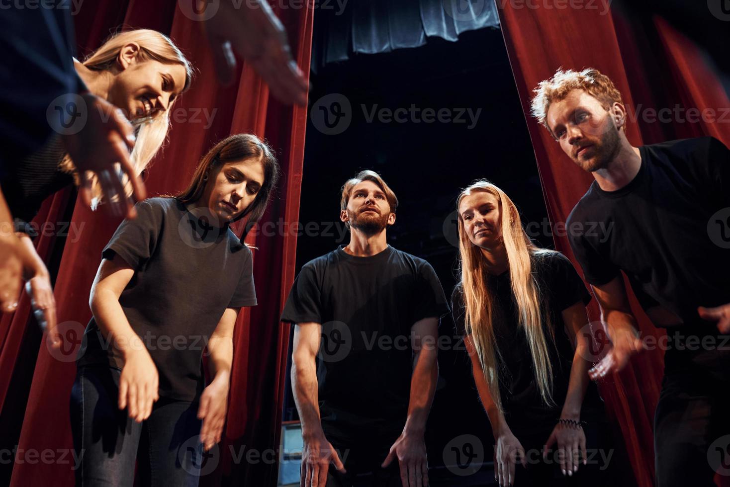 de pie contra las cortinas rojas. grupo de actores con ropa de color oscuro ensayando en el teatro foto