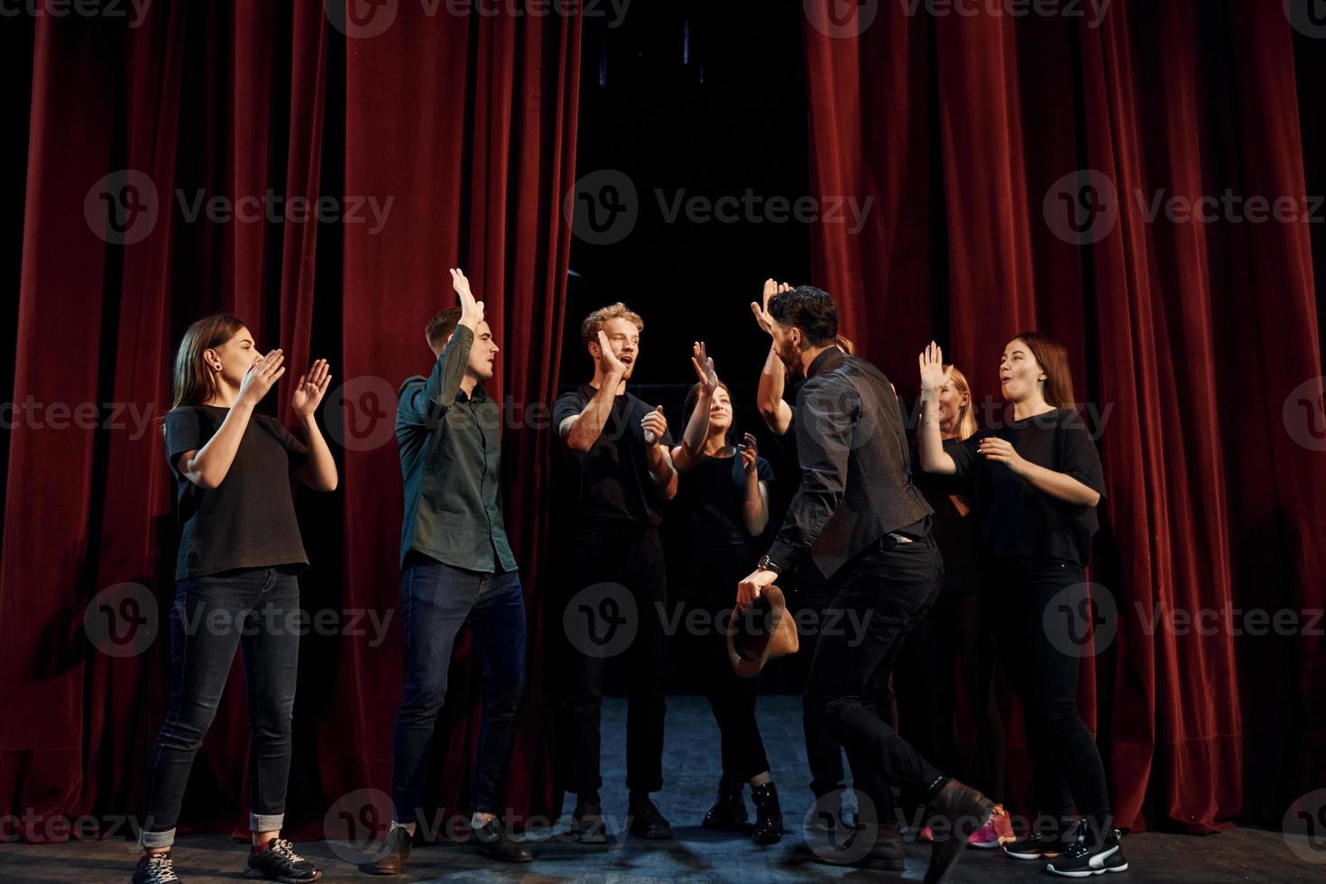 de pie contra las cortinas rojas. grupo de actores con ropa de color oscuro ensayando en el teatro foto