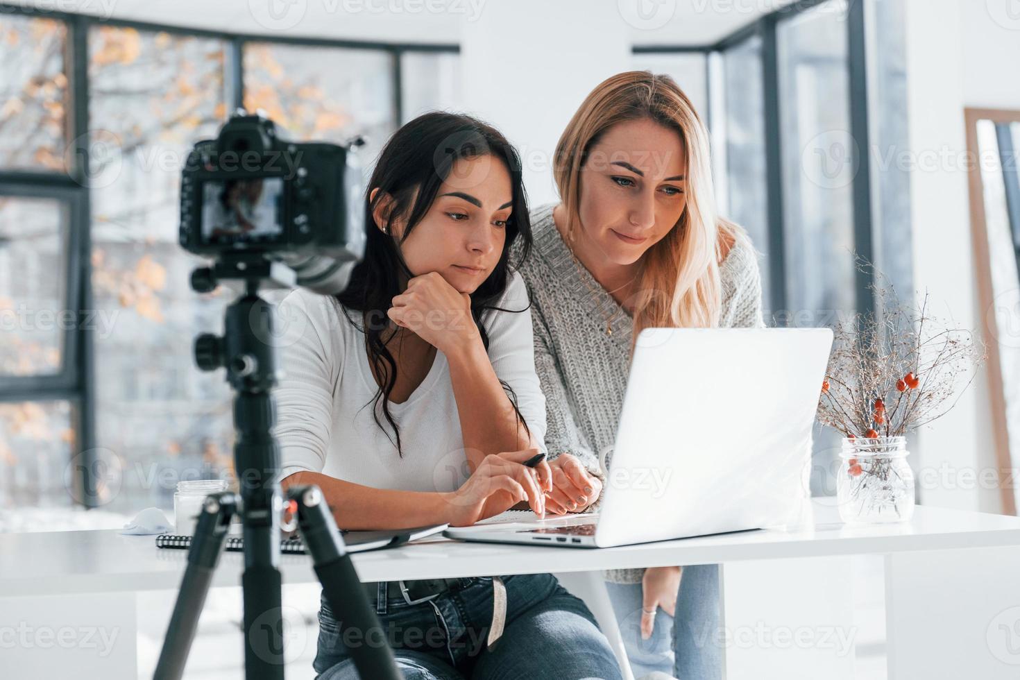 Camera on tripod is recording. Two young female freelancers working indoors in the office with laptop photo