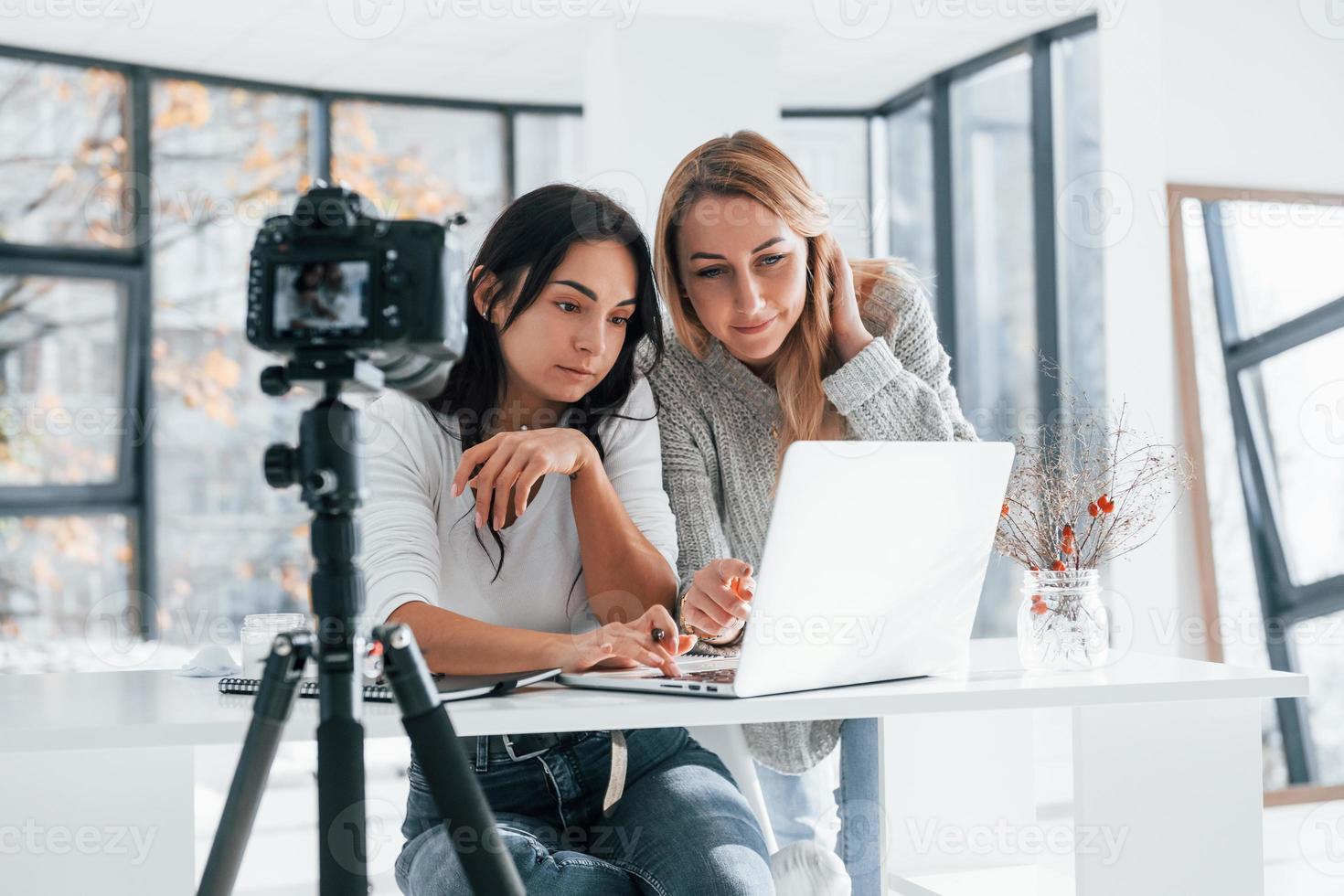 Camera on tripod is recording. Two young female freelancers working indoors in the office with laptop photo