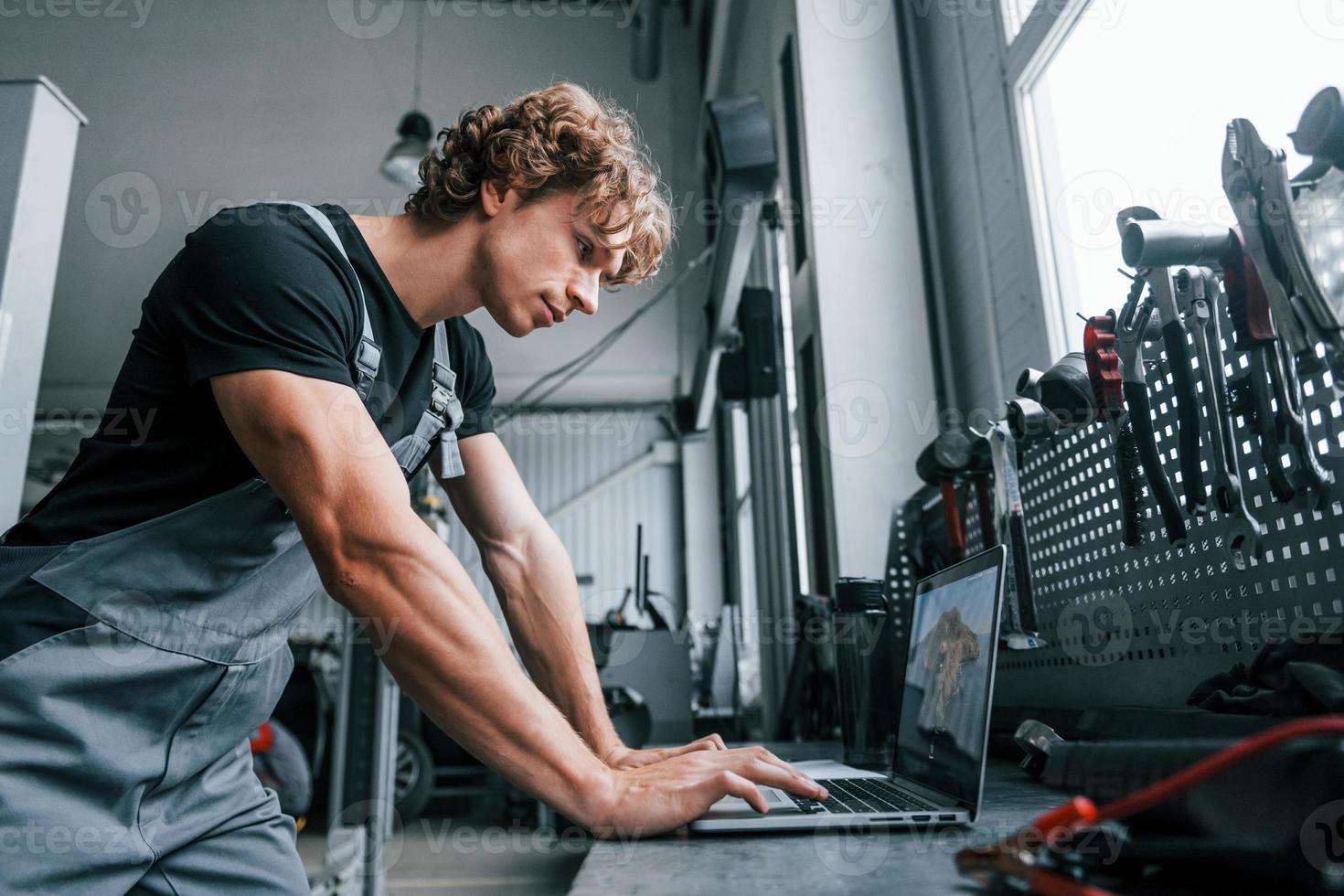 With laptop. Adult man in grey colored uniform works in the automobile salon photo