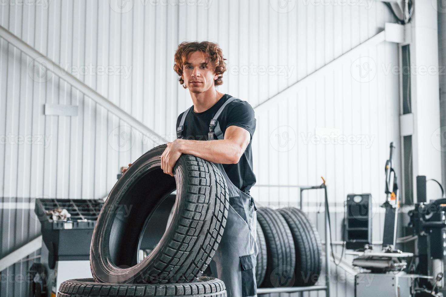 Holds tire. Adult man in grey colored uniform works in the automobile salon photo