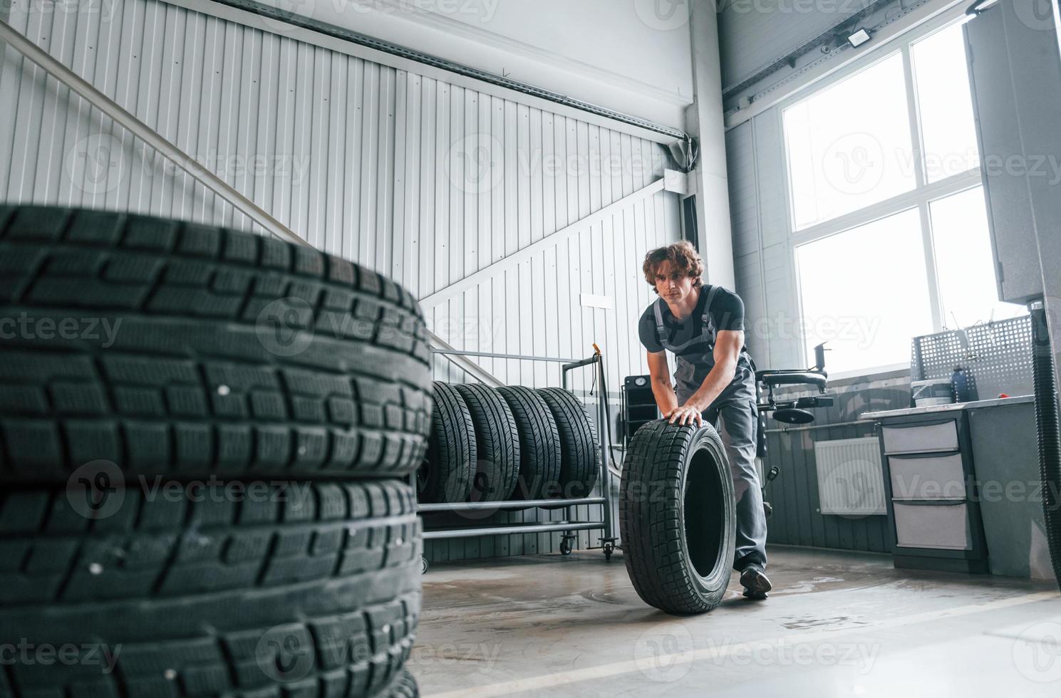 Rolls the tire. Adult man in grey colored uniform works in the automobile salon photo