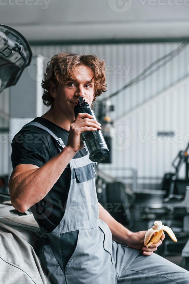 Takes a break. Eats banana and drinks water. Adult man in grey colored uniform works in the automobile salon photo