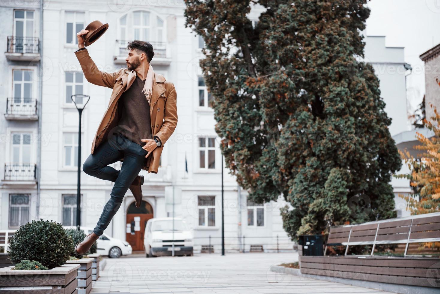 Holds hat and posing. Young male model in fashionable clothes is outdoors in the city at daytime photo