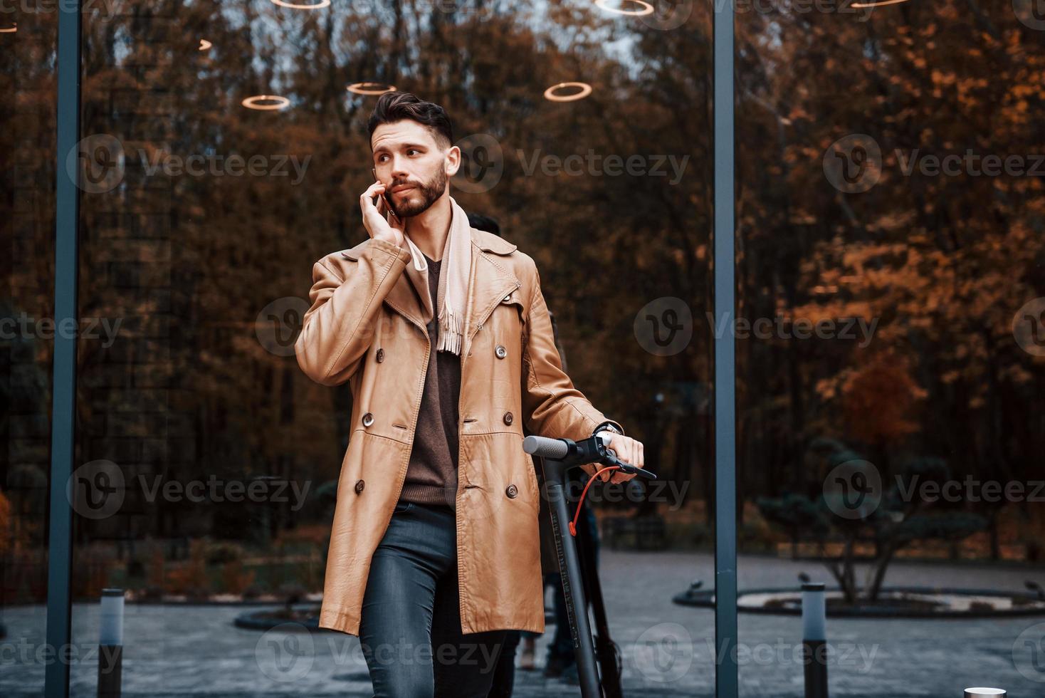 Against building and with electric scooter. Young male model in fashionable clothes is outdoors in the city at daytime photo