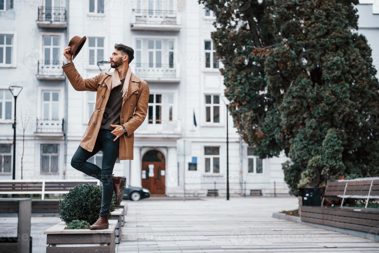 Holds hat and posing. Young male model in fashionable clothes is outdoors in the city at daytime photo