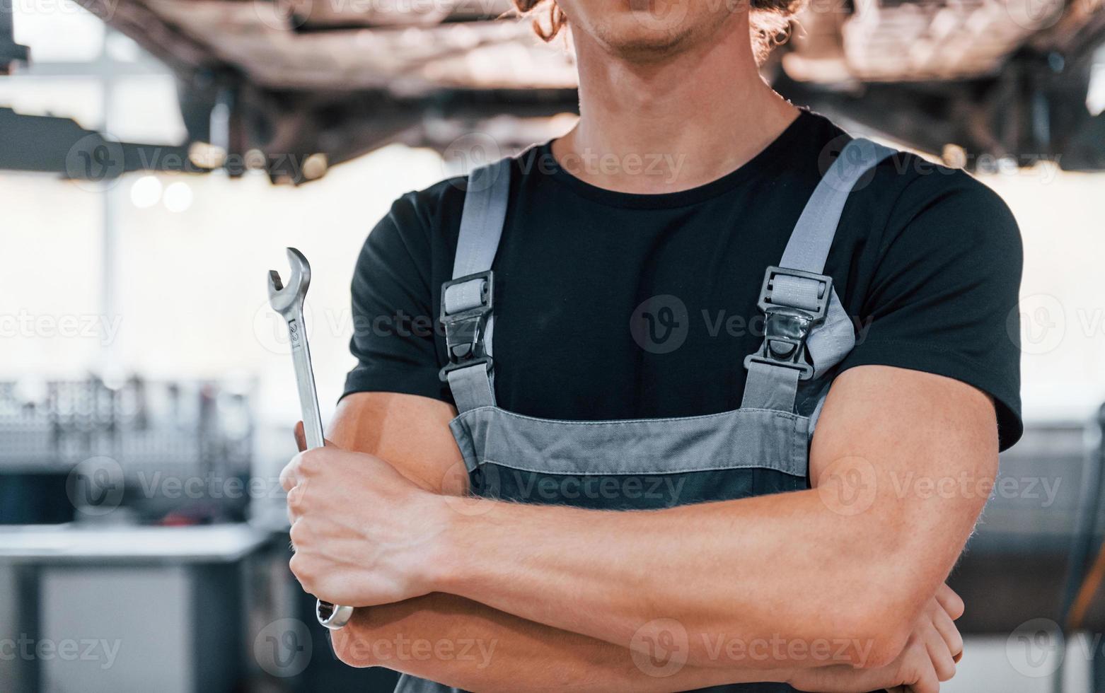 Close up view of guy that holds wrench. Adult man in grey colored uniform works in the automobile salon photo