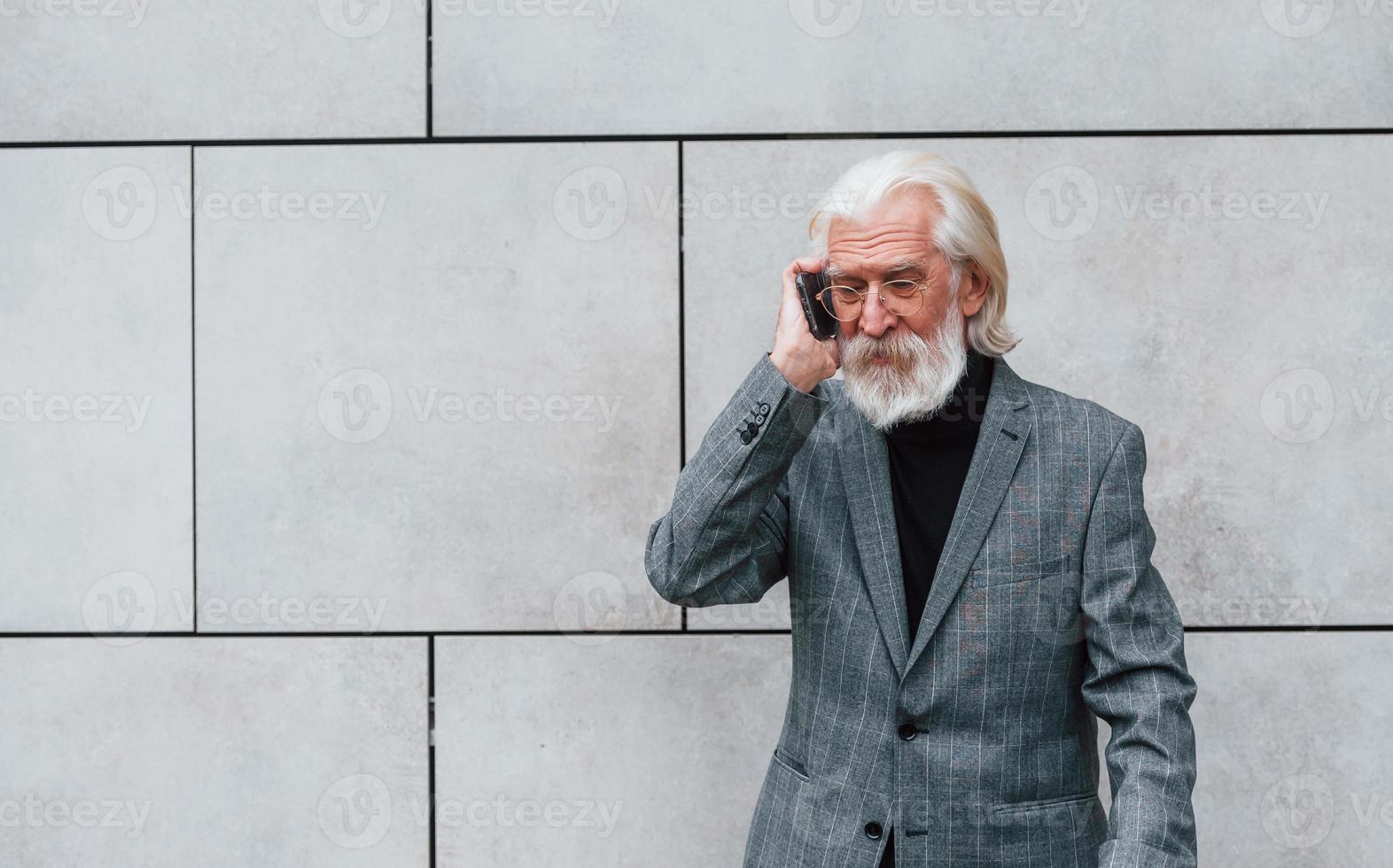 un hombre de negocios de alto nivel con ropa formal, con cabello gris y barba, habla por teléfono al aire libre foto