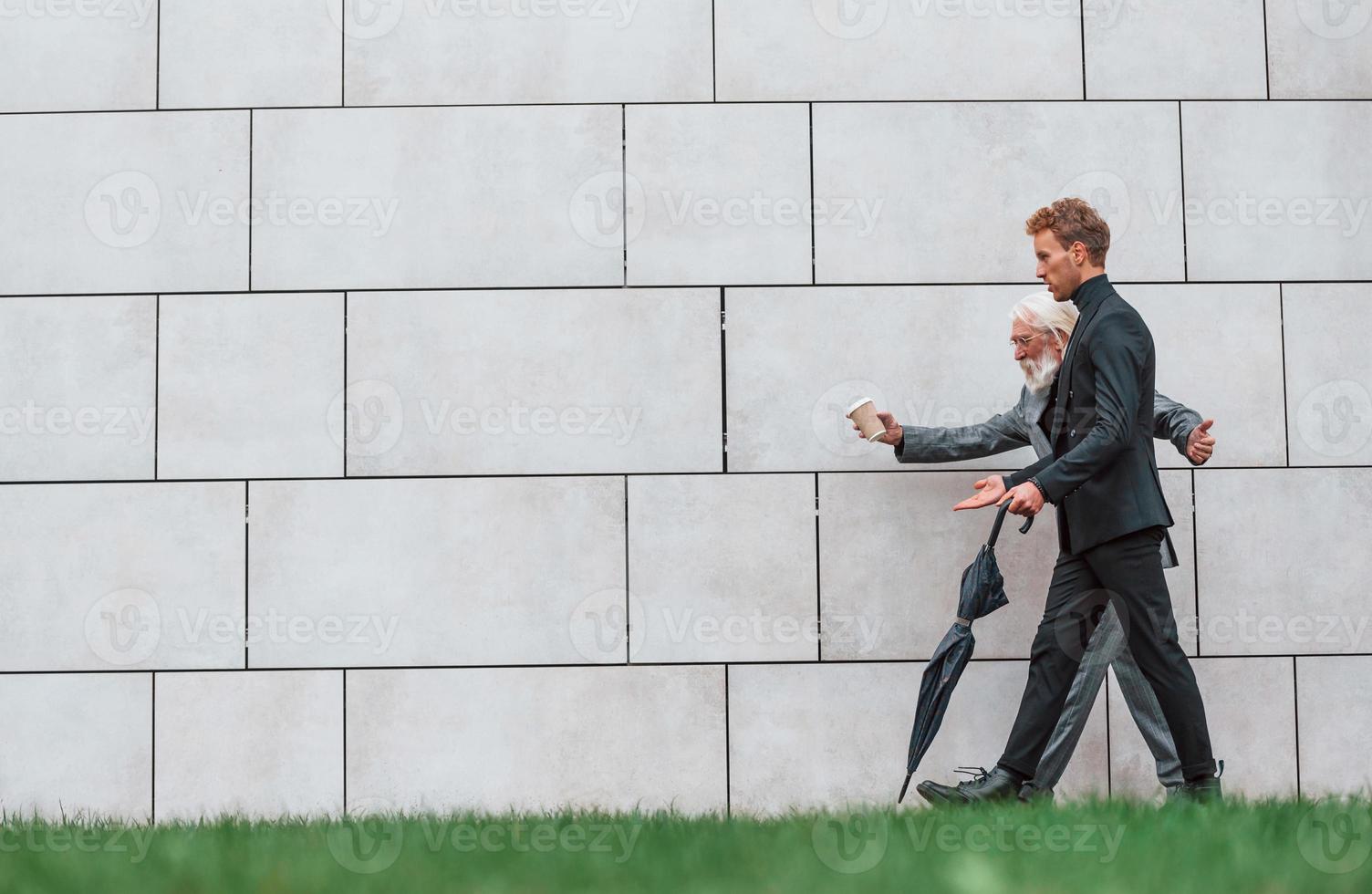 caminando sobre la hierba cerca de la pared. un joven con un anciano vestido con ropa elegante está juntos al aire libre. concepción del negocio foto