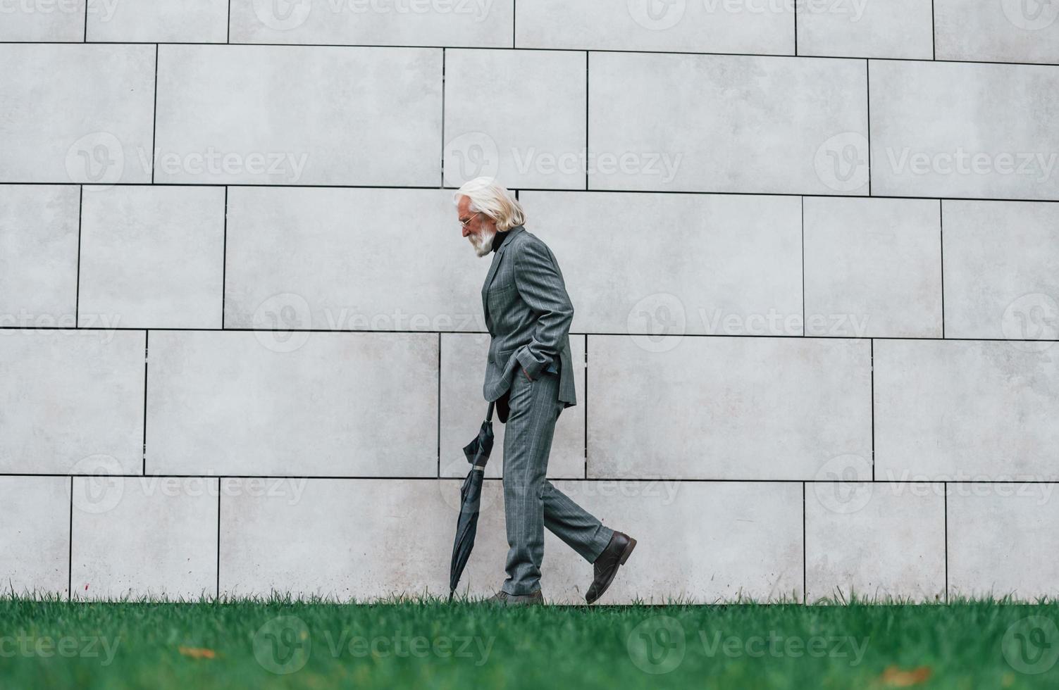 Senior businessman in formal clothes, with grey hair and beard is outdoors walks with umbrella in hand photo