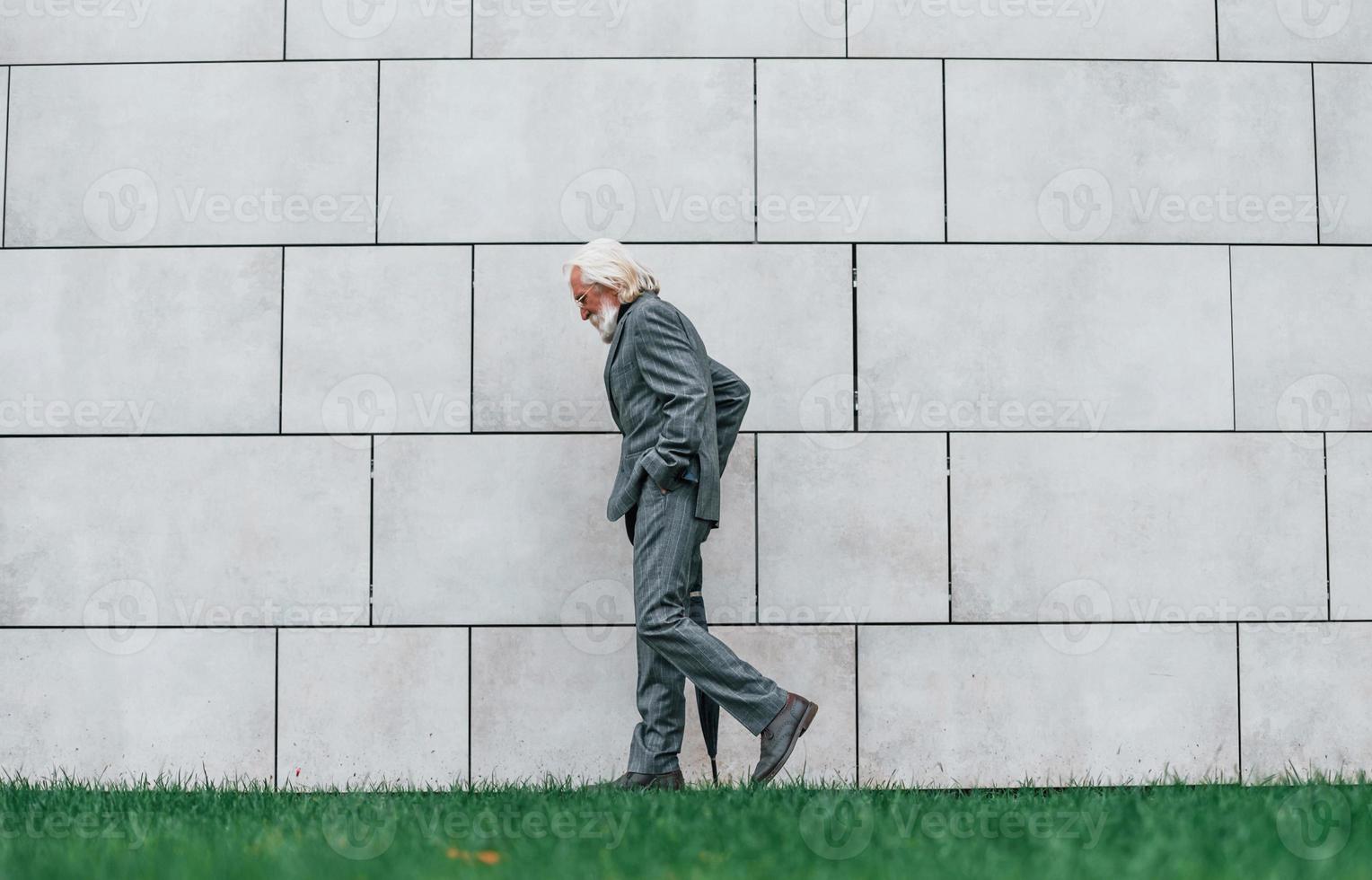 Senior businessman in formal clothes, with grey hair and beard is outdoors walks with umbrella in hand photo