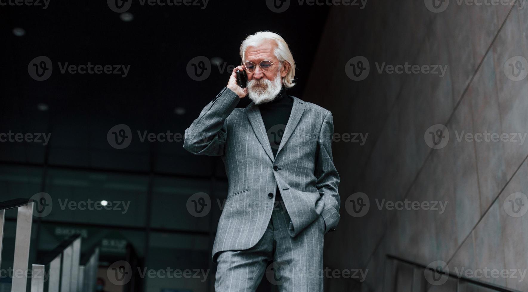 tener una conversación usando el teléfono. hombre de negocios senior en ropa formal, con pelo gris y barba está al aire libre foto