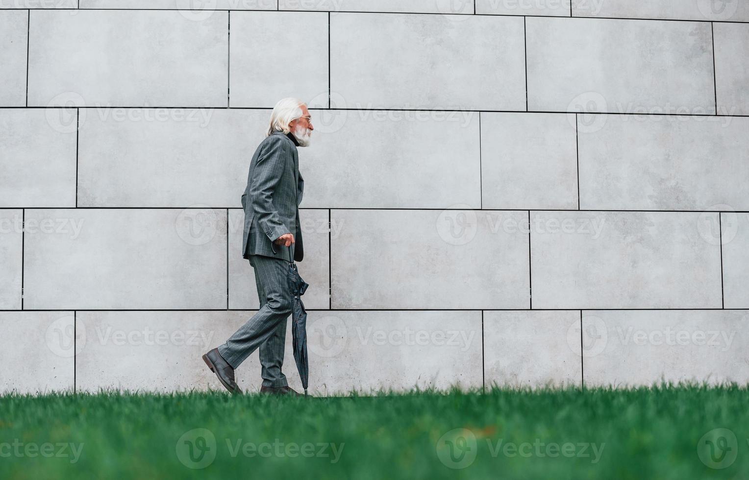 Senior businessman in formal clothes, with grey hair and beard is outdoors walks with umbrella in hand photo