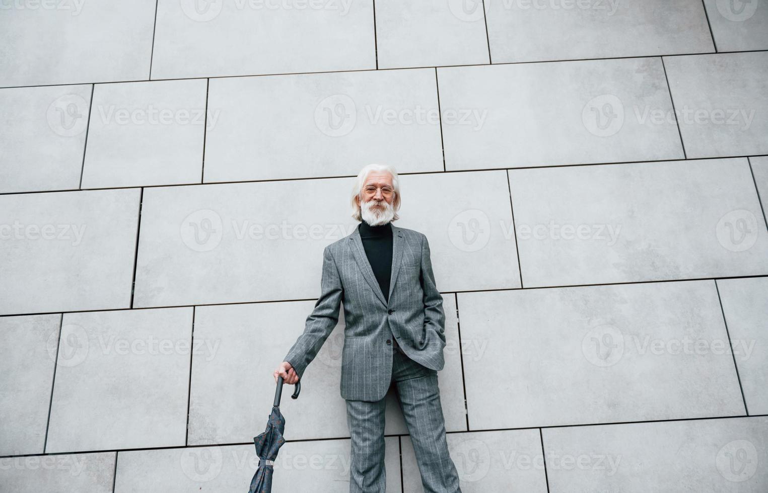sostiene paraguas. hombre de negocios senior en ropa formal, con pelo gris y barba está al aire libre foto