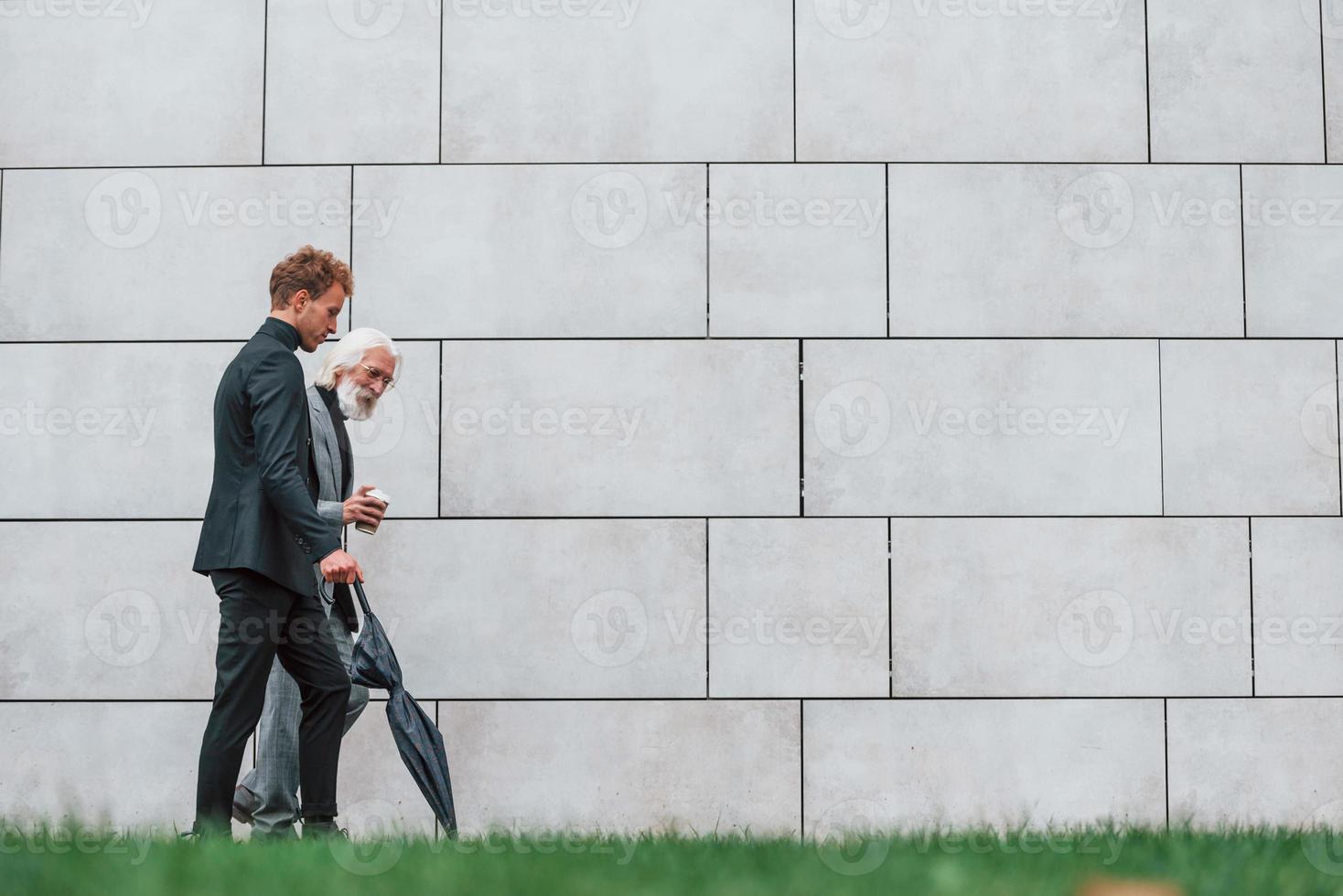 Walking on grass near wall. Young guy with senior man in elegant clothes is outdoors together. Conception of business photo