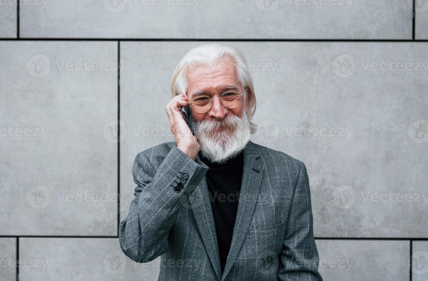 un hombre de negocios de alto nivel con ropa formal, con cabello gris y barba, habla por teléfono al aire libre foto