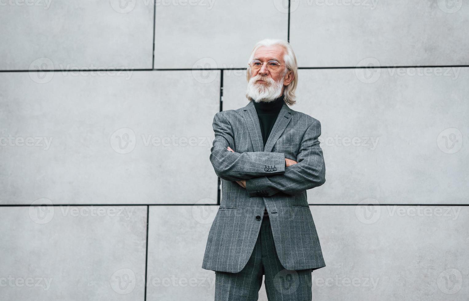 Senior businessman in formal clothes, with grey hair and beard is outdoors stands against the wall photo