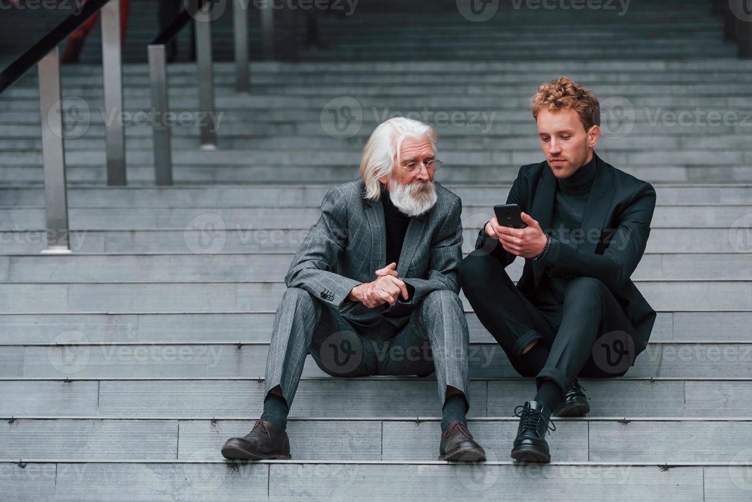 Sitting with phone. Young guy with senior man in elegant clothes is outdoors together. Conception of business photo