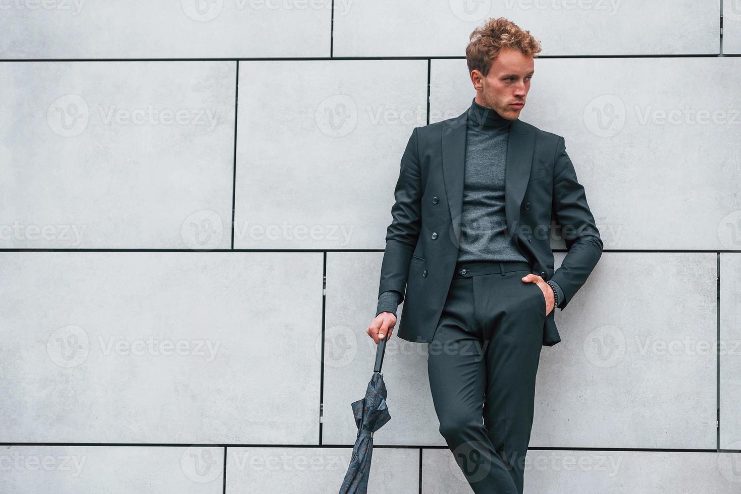 joven en ropa formal con paraguas de pie cerca de la pared al aire libre con paraguas foto