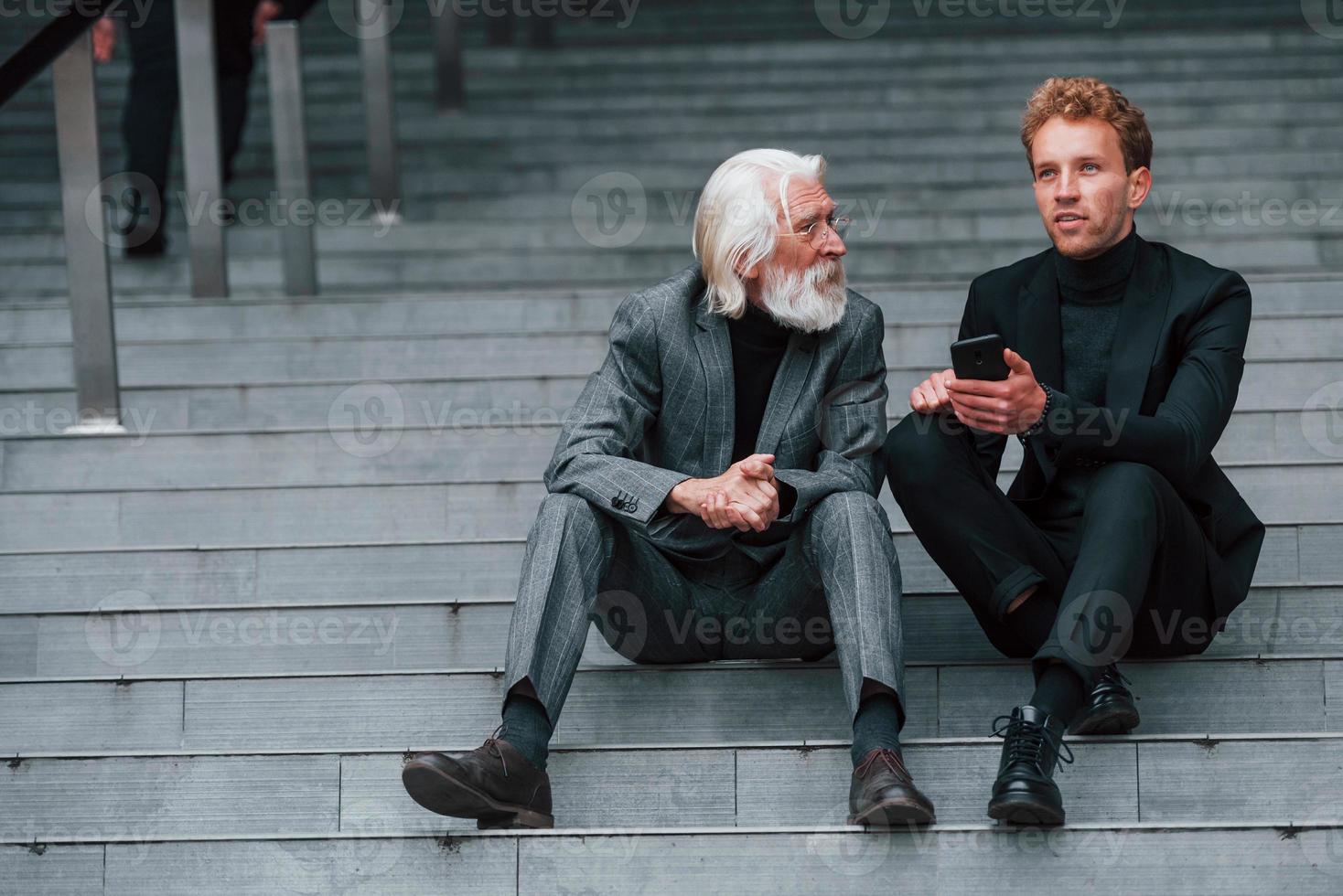 Sitting with phone. Young guy with senior man in elegant clothes is outdoors together. Conception of business photo