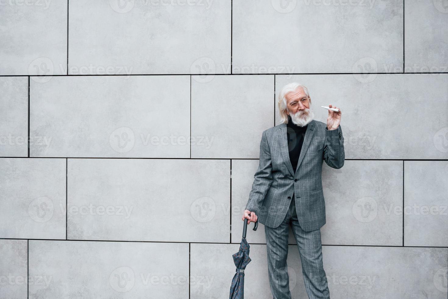 fumar cigarrillo electrónico. hombre de negocios senior en ropa formal, con pelo gris y barba está al aire libre foto