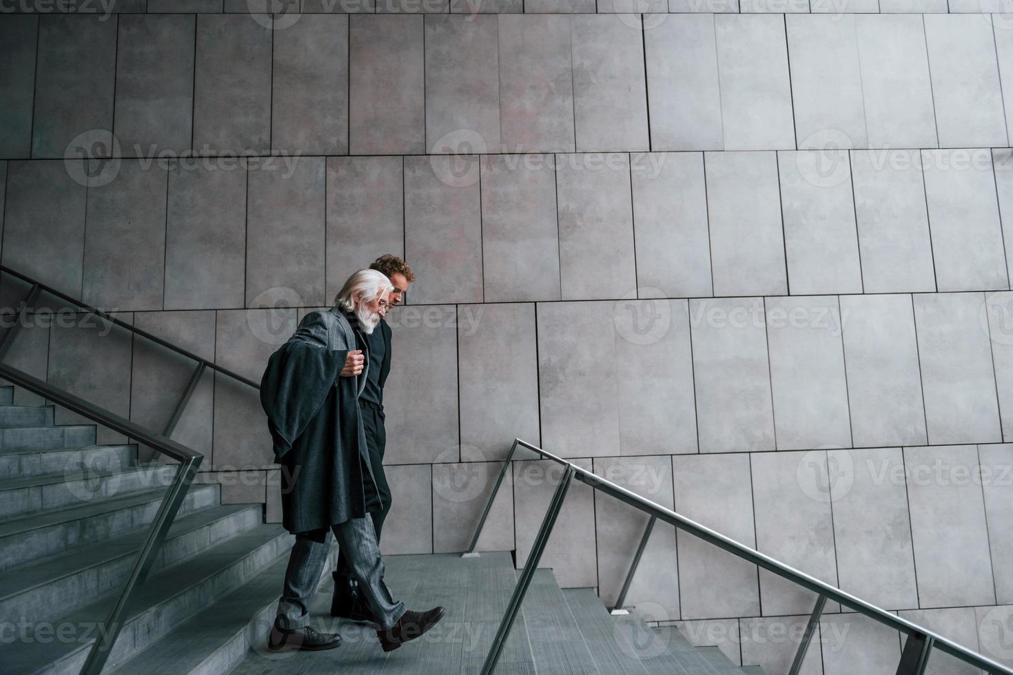 Walking on the stairs. Young guy with senior man in elegant clothes is outdoors together. Conception of business photo