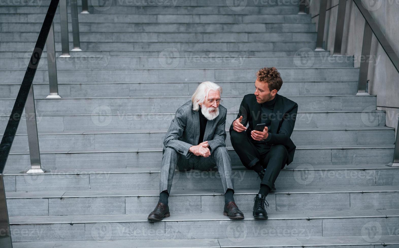 Sitting with phone. Young guy with senior man in elegant clothes is outdoors together. Conception of business photo