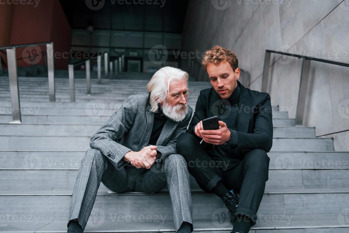 Sitting with phone. Young guy with senior man in elegant clothes is outdoors together. Conception of business photo