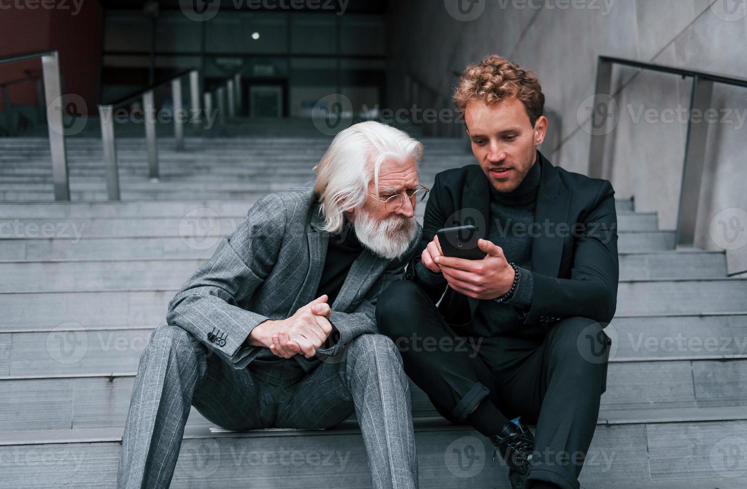 Sitting with phone. Young guy with senior man in elegant clothes is outdoors together. Conception of business photo