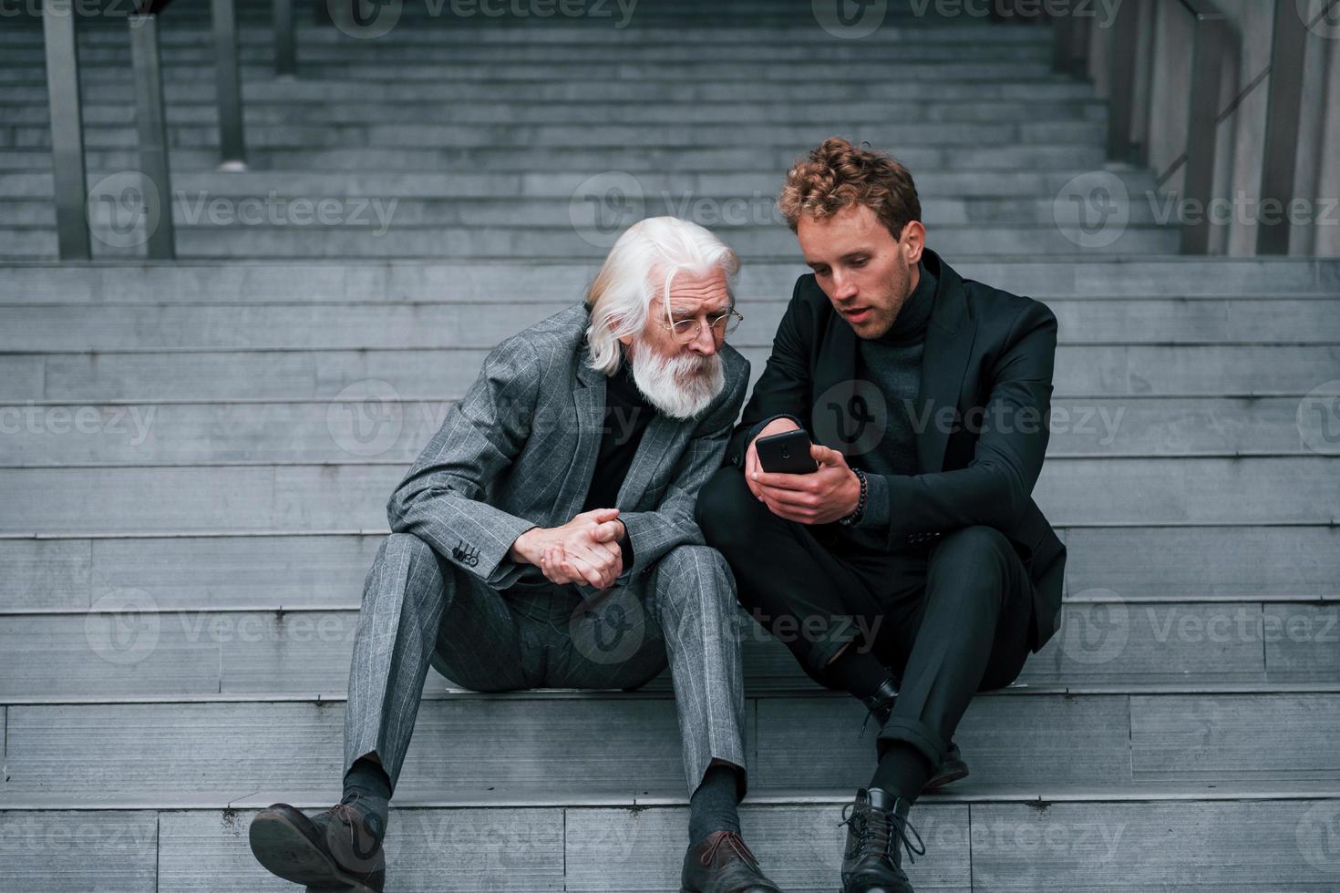 Sitting with phone. Young guy with senior man in elegant clothes is outdoors together. Conception of business photo