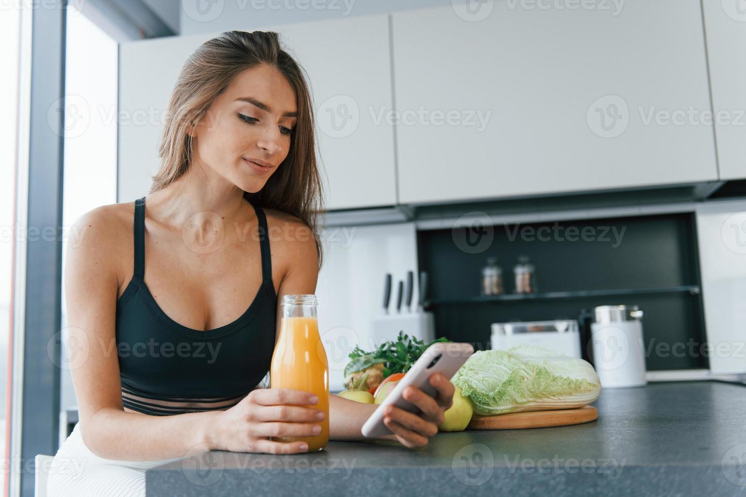 Uses smartphone. Young european woman is indoors at kitchen indoors with healthy food photo