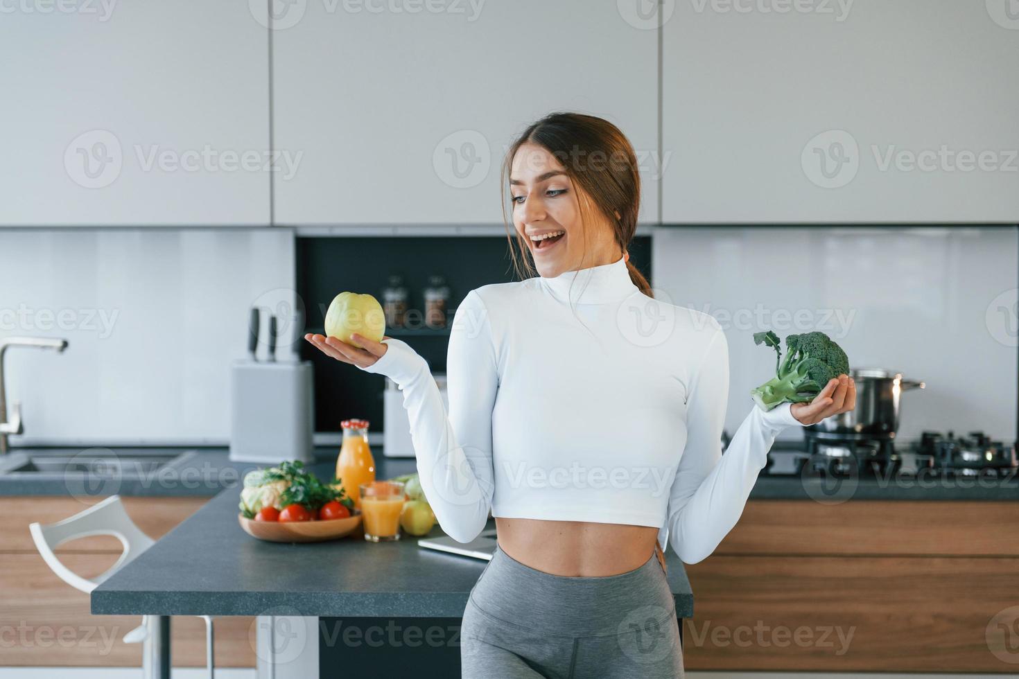 Poses for a camera with vegetables. Young european woman is indoors at kitchen indoors with healthy food photo