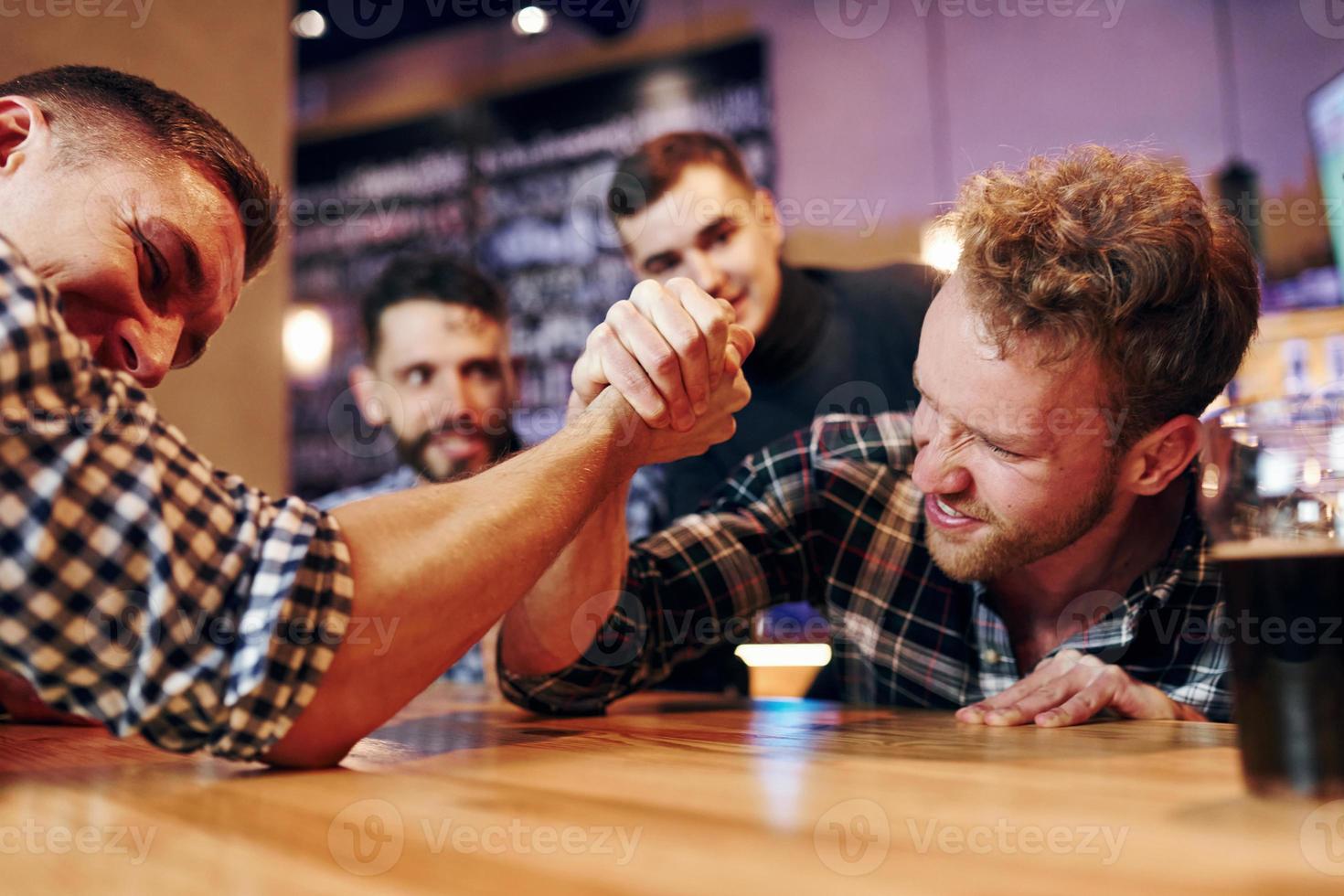 Friends plays arm wrestling. Group of people together indoors in the pub have fun at weekend time photo