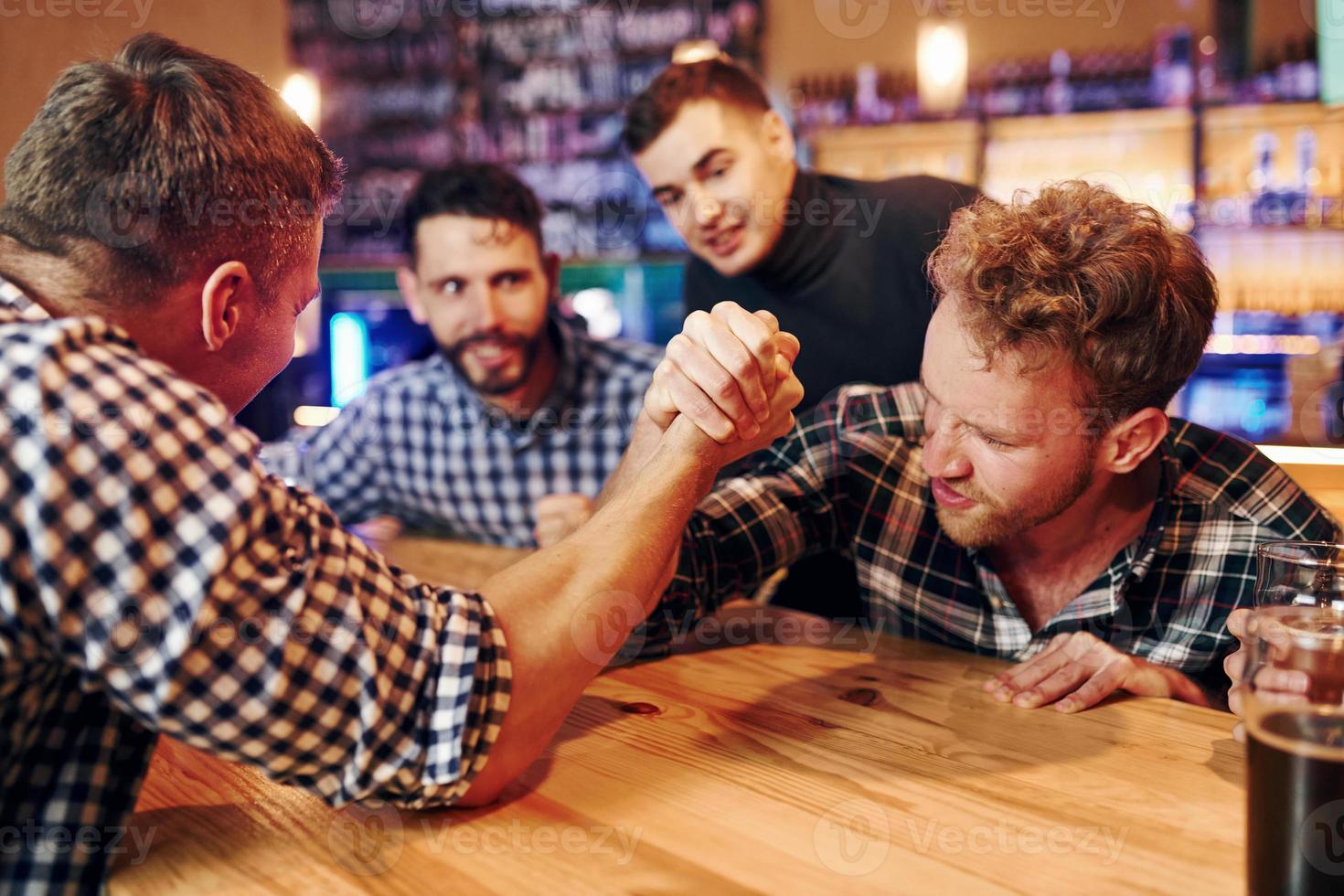 Friends plays arm wrestling. Group of people together indoors in the pub have fun at weekend time photo