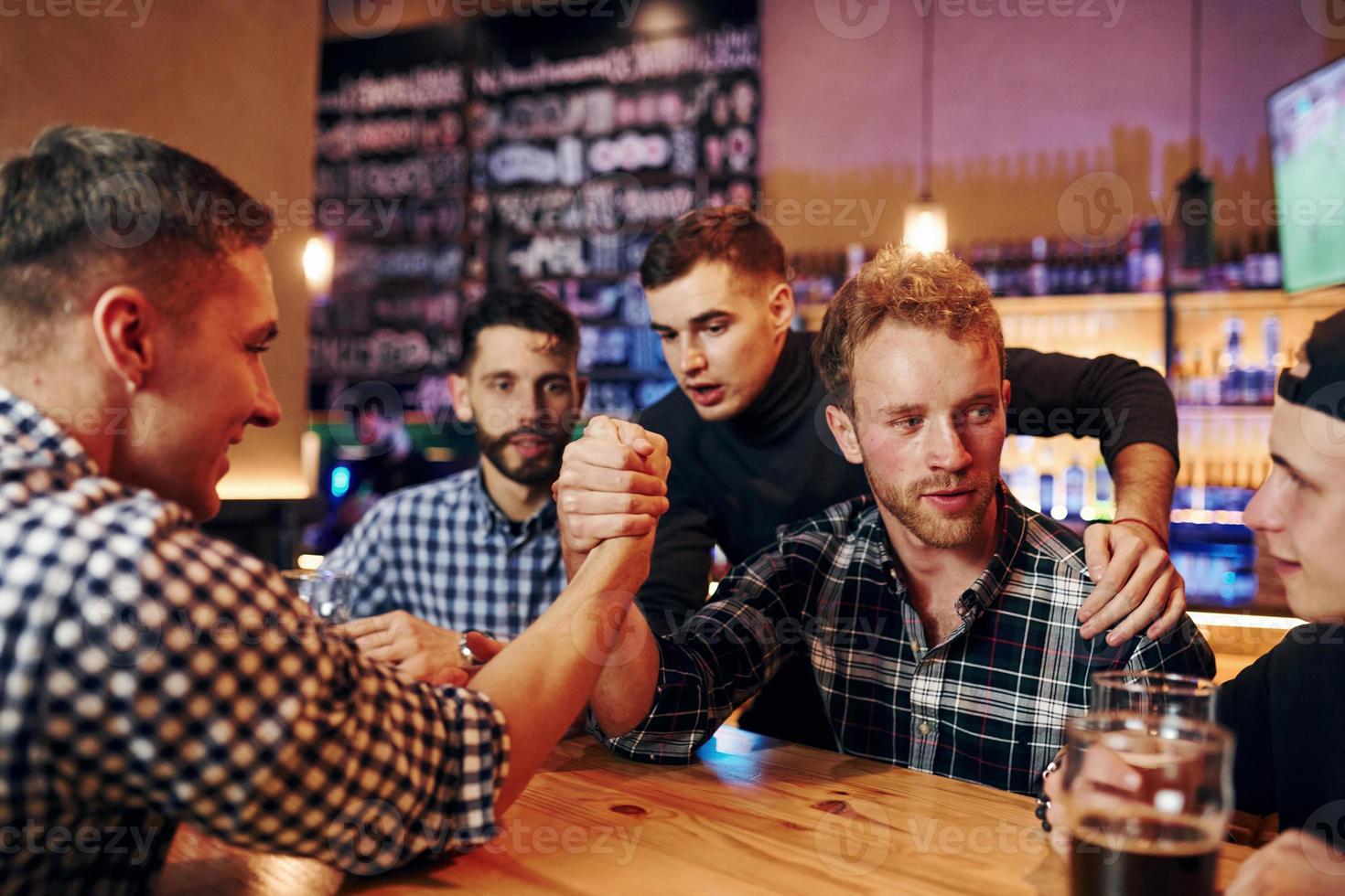 Friends plays arm wrestling. Group of people together indoors in the pub have fun at weekend time photo