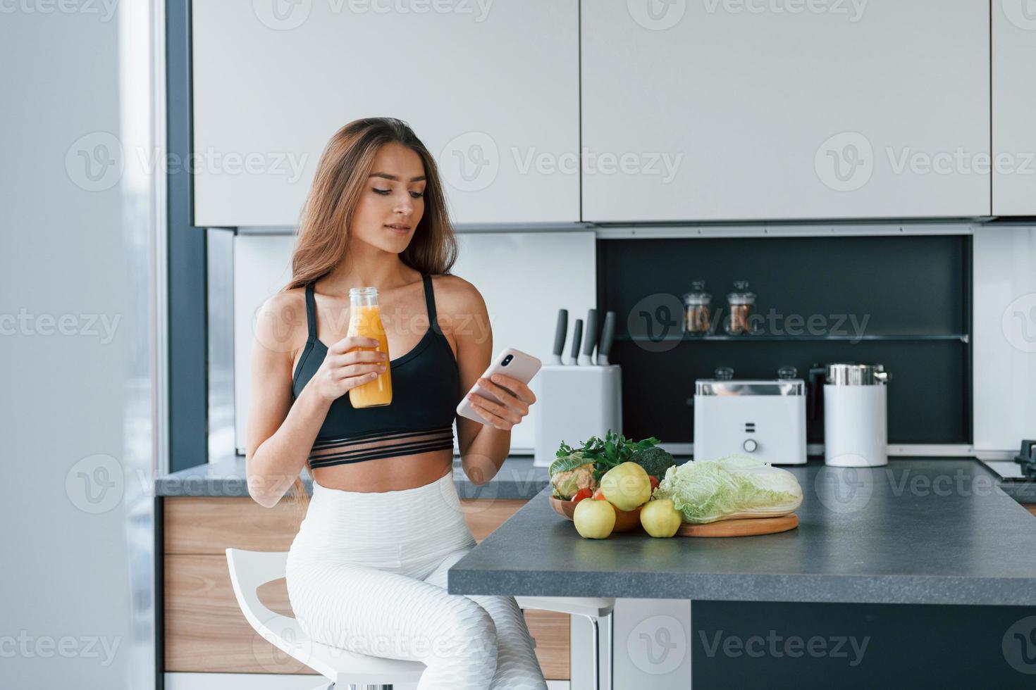 Uses smartphone. Young european woman is indoors at kitchen indoors with healthy food photo