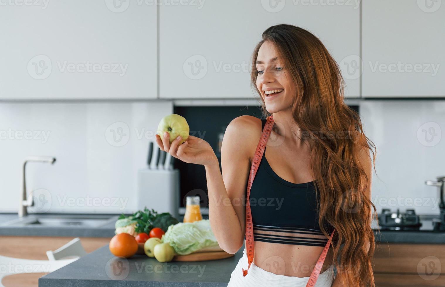 Holds apple in hand. Young european woman is indoors at kitchen indoors with healthy food photo