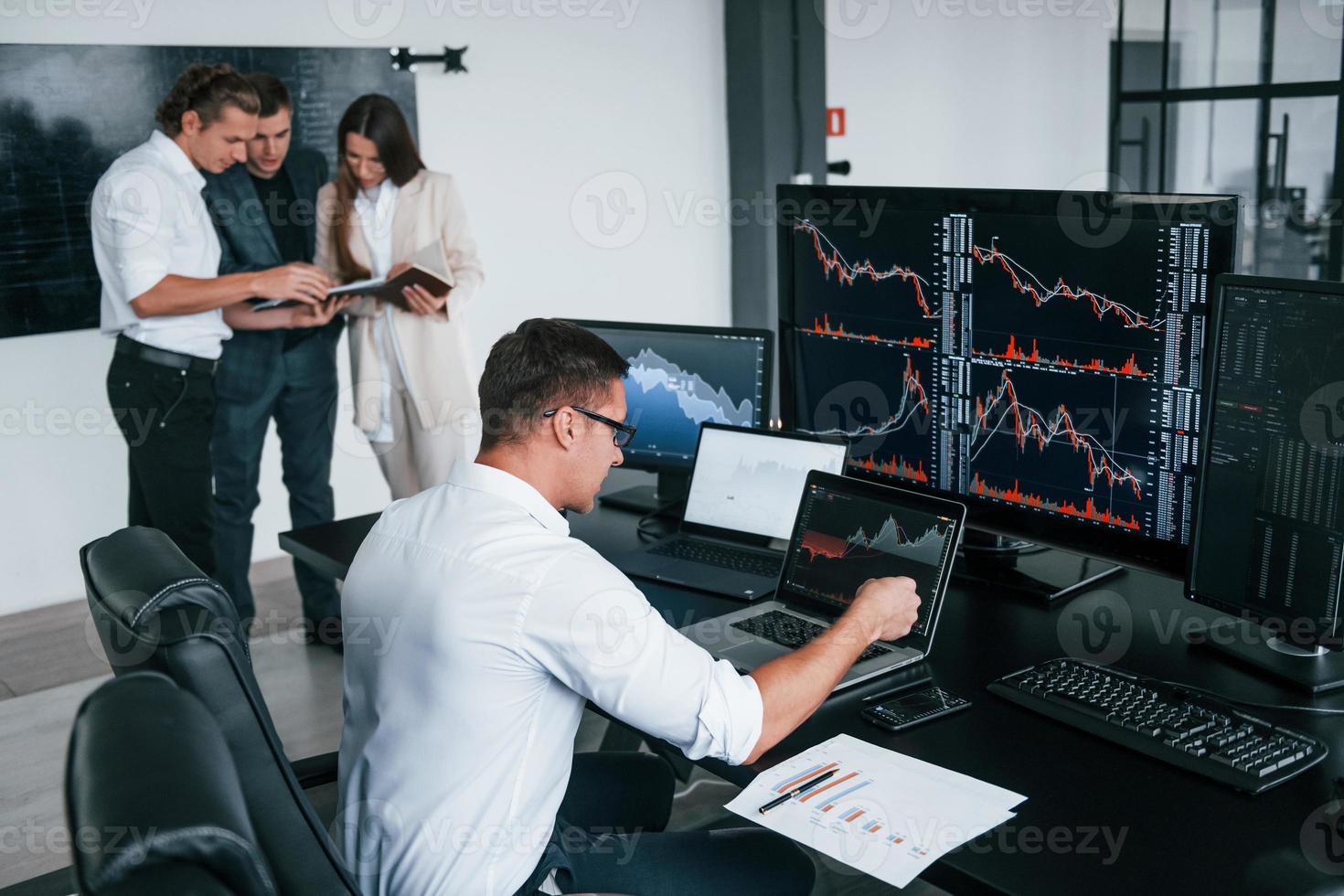 Man uses laptop. Team of stockbrokers works in modern office with many display screens photo