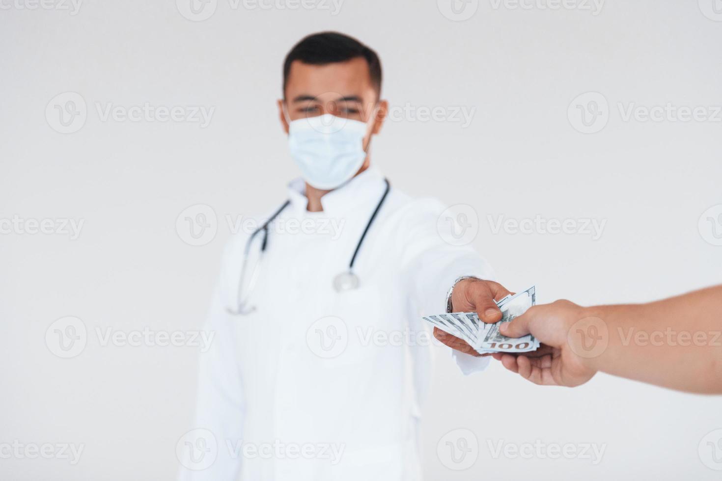 Medic holds money. Young handsome man standing indoors against white background photo