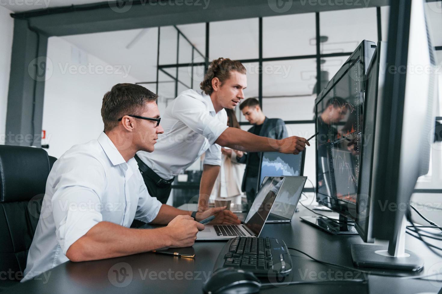 Guy in glasses concentrated at work. Team of stockbrokers is in modern office with many display screens photo