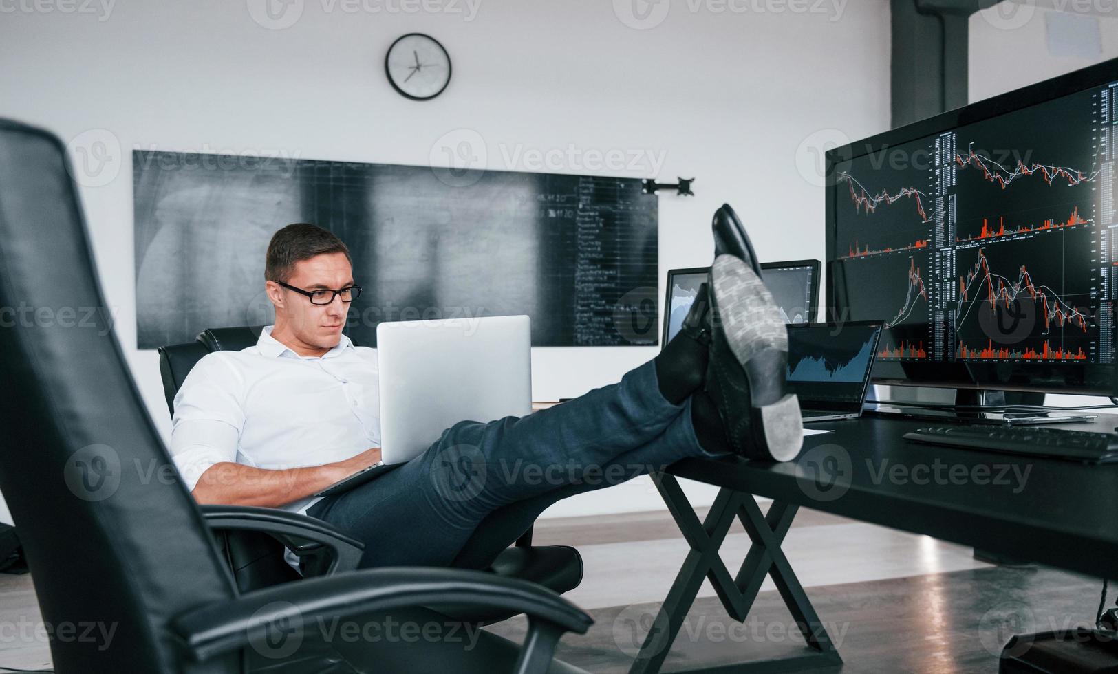 Sits on the chair. Young male stock brocker works indoors in the office. Many of displays photo