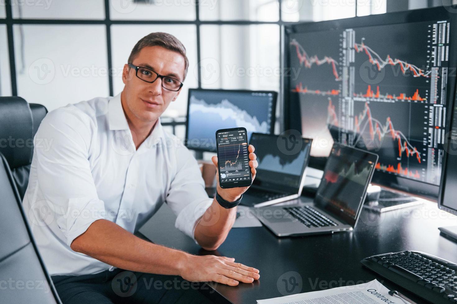 Holds phone. Young male stock brocker works indoors in the office. Many of displays photo