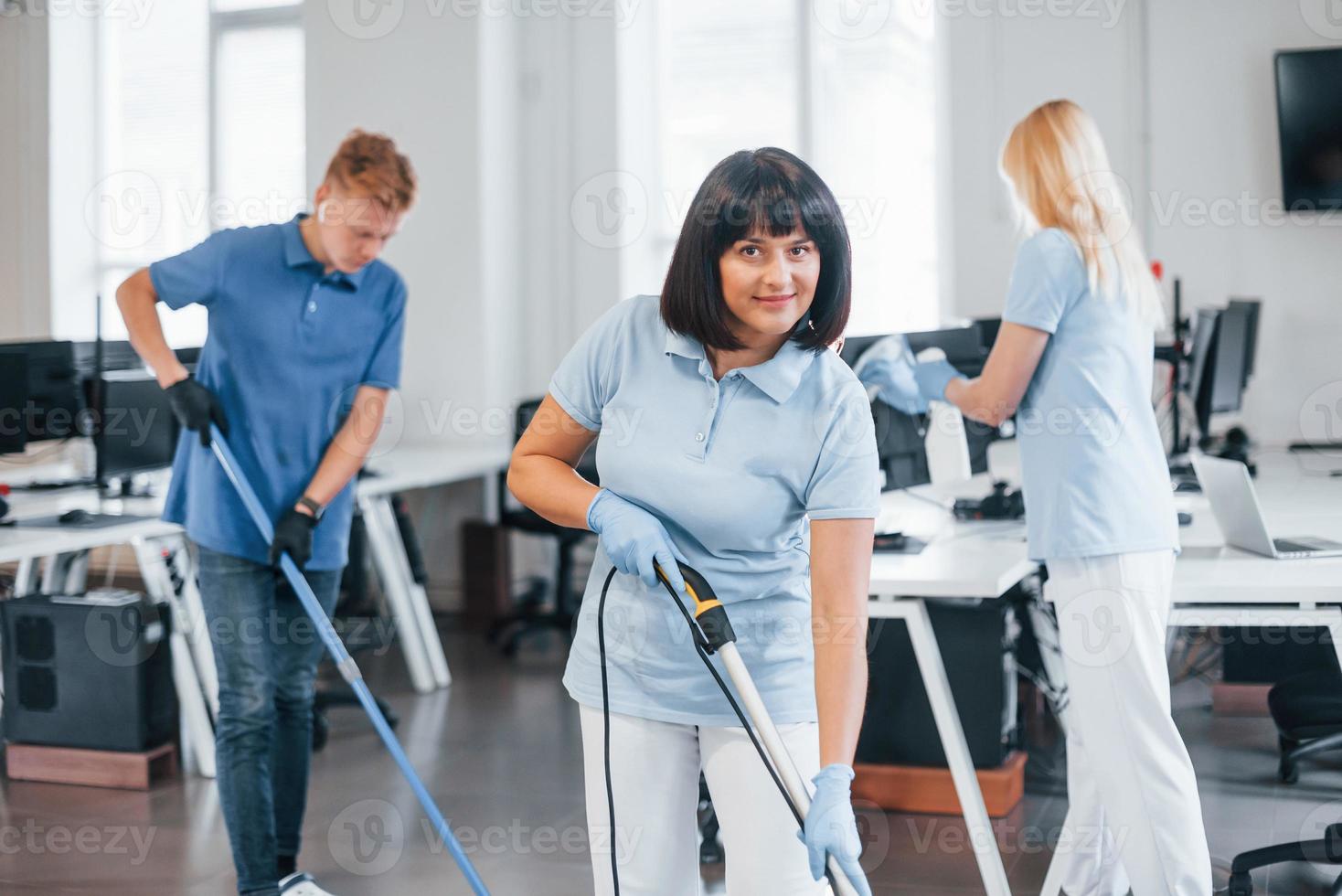 Woman uses vacuum cleaner. Group of workers clean modern office together at daytime photo