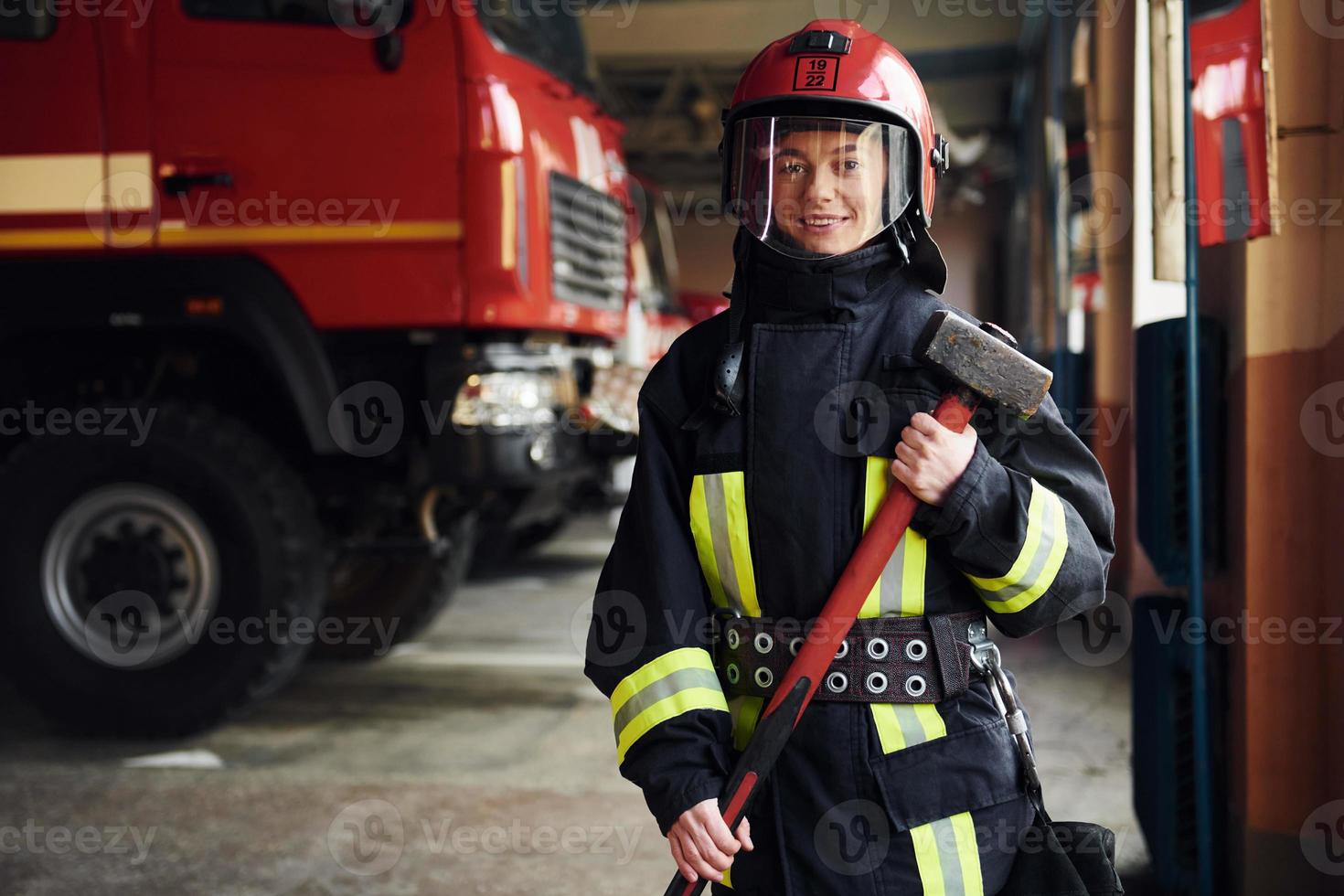 With hammer in hands. Female firefighter in protective uniform standing near truck photo